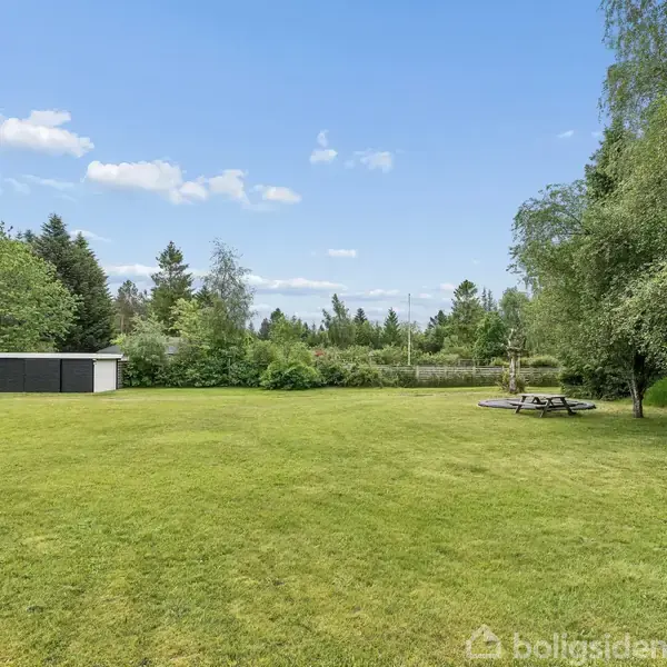 Græsplæne med et picnicbord under et træ. Et sort hus med rødt tag og en mindre bygning ses til venstre. Baggrunden er domineret af grønne træer under en blå himmel.