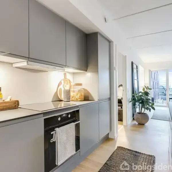 Køkken med grå skabe har urtepotter og krydderier på bordet. I baggrunden er en stue med store vinduer og planter. Lys, moderne indretning præger hele rummet.