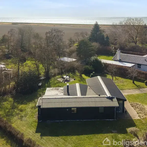 Et sort hus med gråtag står i en grøn have. Ombordliggende er andre huse, træer og buske. I baggrunden ses en strand og havet.