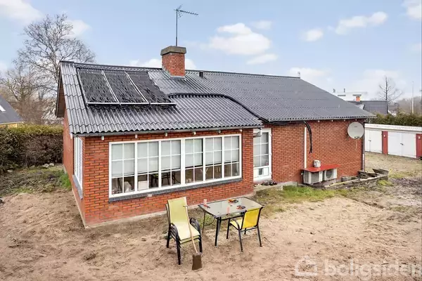 Rødt murstenshus med sort tag og solpaneler. Foran huset står to stole og et bord på en bar jordplæne. I baggrunden ses en hæk og en garage.