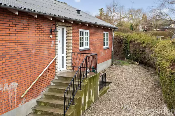 Rød murstenshus med hvid dør og vinduer, placeret ved en grusbelagt indkørsel. Trappen op til døren har sort gelænder. Omgivet af hæk og træer i en have.