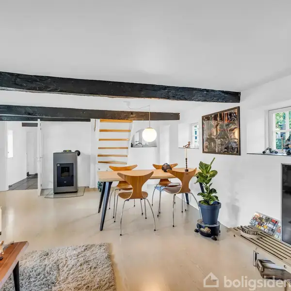 Spisestue med træbord og stole under en moderne lampe, omgivet af hvide vægge, synlige bjælker og en grøn plante. Pejs og trapper i baggrunden. Hyggelig, åben atmosfære.