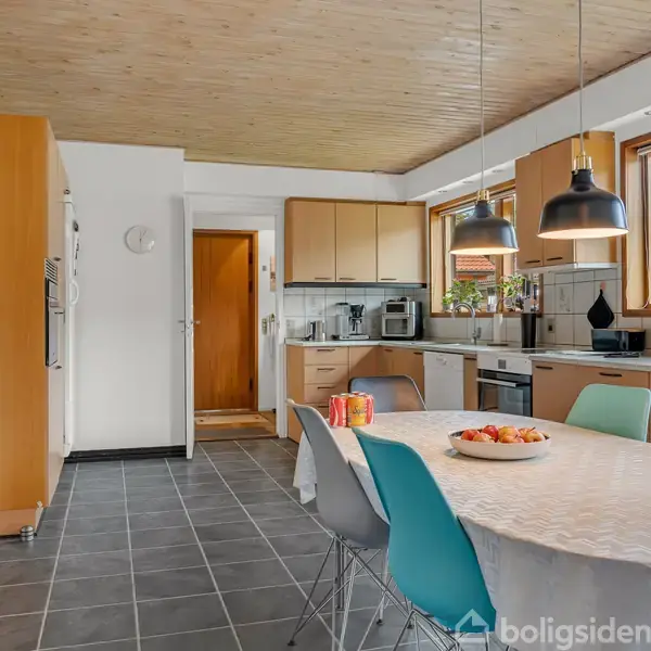 Køkken med spisebord dækket med en dug og dekoreret med en frugtskål og blomster. Flere stole omgiver bordet, og moderne lamper hænger over. Store vinduer giver naturligt lys.