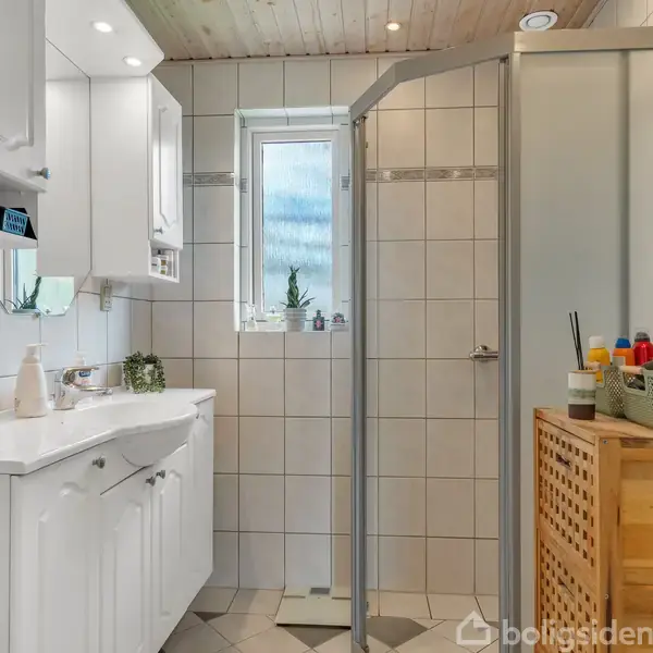 Badeværelse med hvidt skabsarrangement og vask, brusekabine i glas, flisevægge, og et lille vindue. Ved siden af bruseren står en trækommode med toiletartikler på toppen.