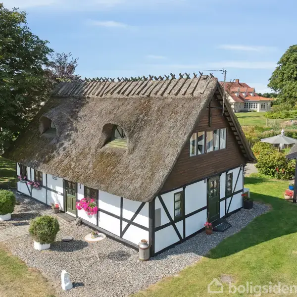 Et bindingsværkshus med stråtag står roligt. Det har små blomster på vindueskarmen og et negligeret græsareal omgivet af træer og en nærliggende bygning.