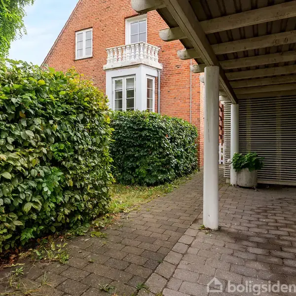 Carport med åben konstruktion står ved siden af et rødt murstenshus. Huset har en lille altan med gelænder. Omgivet af grønne buske og flisebelagt gangsti.