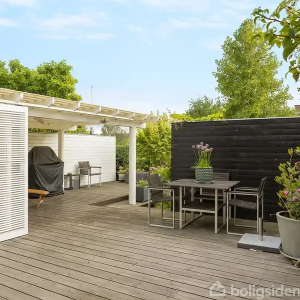 Terrasseområde med træmøbler og planter, omgivet af træhegn og buske. En grill står under et overdækket område med åbent, lyst loft, der giver et luftigt indtryk.
