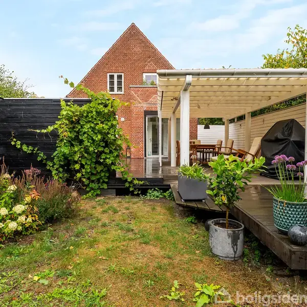 Et træterrasseområde foran et rødt murstenshus har havemøbler under en hvid pergola. Omkranset af frodige planter og buske, skaber det en hyggelig atmosfære.