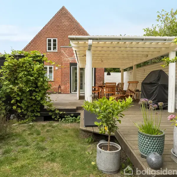 En rød murstensbygning med terrasse i en have. Terrassen har et overdækket spiseområde med træbord og grill. Omgivet af grønne planter og blomster i krukker.