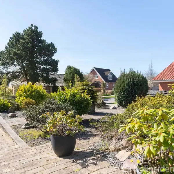 En velplejet have med forskellige buske og træer omgivet af stier. I baggrunden ses et rødt hus med tegltag og flere træer. Det er solrigt og klart vejr.