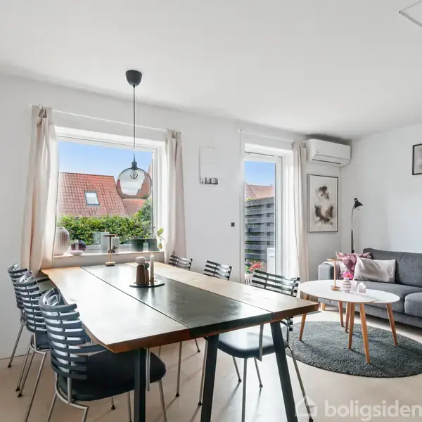 En lys stue med et stort spisebord og stole til venstre, ved siden af en sofagruppe med gråt tæppe og sofabord til højre. Store vinduer giver udsigt til hustage.