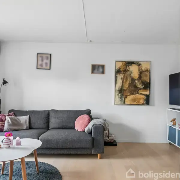 Grå sofa med puder står op ad en hvid væg med kunstværker. Til venstre er der en gulvlampe og et vindue. Rundt sofabord med små pynteting. Reol med bøger i baggrunden.