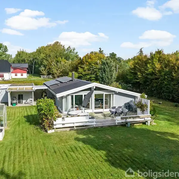 Et hvidt hus med store vinduer står i en velplejet have med en terrasse. Ved siden af huset er der et drivhus. Træer og buske omgiver grunden under en blå himmel.