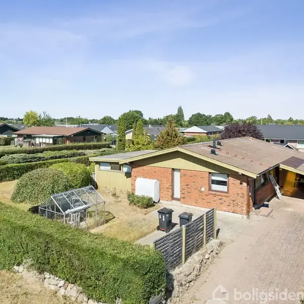 Et enfamiliehus af mursten står i et villakvarter. Foran huset er en carport, en lille have med et drivhus, og alt er omgivet af hække.