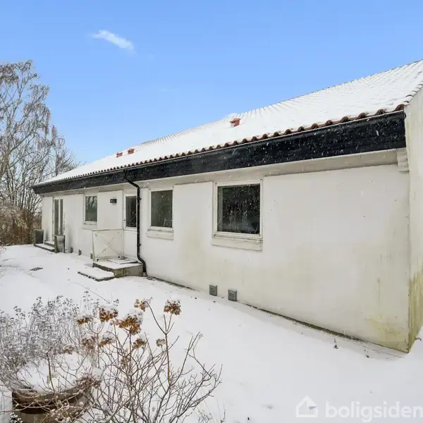 Hus i hvidt, sneklædt landskab; det står stille om vinteren med sne på taget og jorden. Sne dækker også buske og træer i baggrunden under en blå himmel.