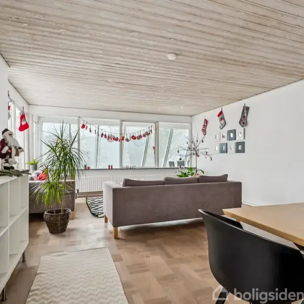 En stue med sofaer er julepyntet med sokker og guirlander. Store vinduer lukker lys ind i rummet, hvor en spisebord med stole står ved siden af et reolsystem.