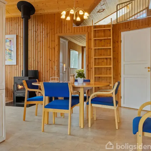 Et rundt træbord med blåpolstrede stole omkring står i en hyggelig stue. Baggrunden har en brændeovn, træbeklædte vægge og loft samt en stige op til en hems.