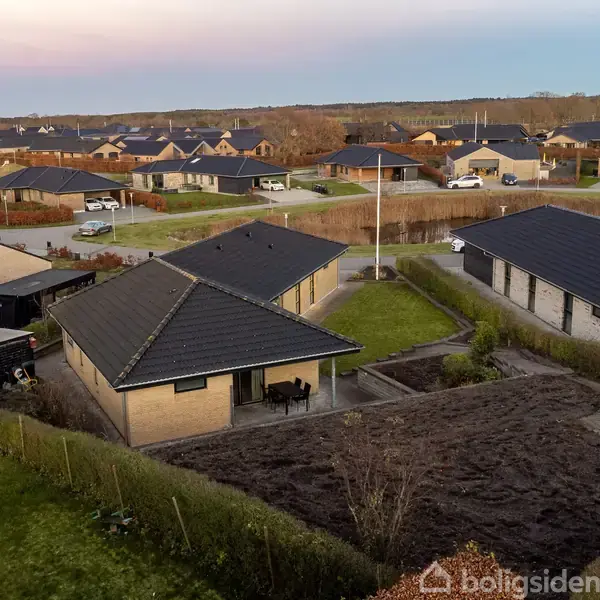 Et enetages hus med skråt tag er omgivet af en have i et boligkvarter. Flere lignende huse kan ses i nabolaget, der består af stille gader og grønne arealer.