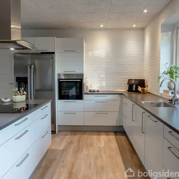 Et moderne køkken med hvide skabe og grå bordplader. Der er en indbygget ovn og et stort køleskab. Bordpladen har en vask og en kaffemaskine. Trægulv og vindue til højre.