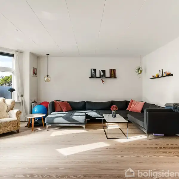 En stor, lys stue har en grå hjørnesofa med farvede puder. Aktion: Sofaen står mod væggen. Kontekst: Rummet har trægulv, en lænestol, vinduer, og udsmykkede hylder.
