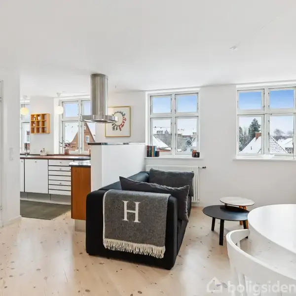 En stue med en sort sofa med tæppe, hvor der står "H". Mod et lyst træbord er der en åben forbindelse til et køkken med hvide skabsfronter og flere vinduer i loftshøjde.