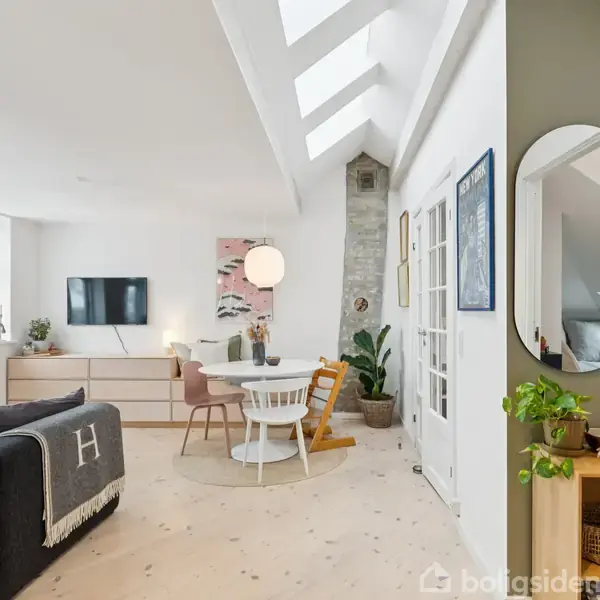 En lys stue med spisebord og stole, sofa med tæppe, væg-TV og planter. Store vinduer og ovenlys vinduer skaber et rummeligt indtryk. Der er også et spejl og dekorative elementer.