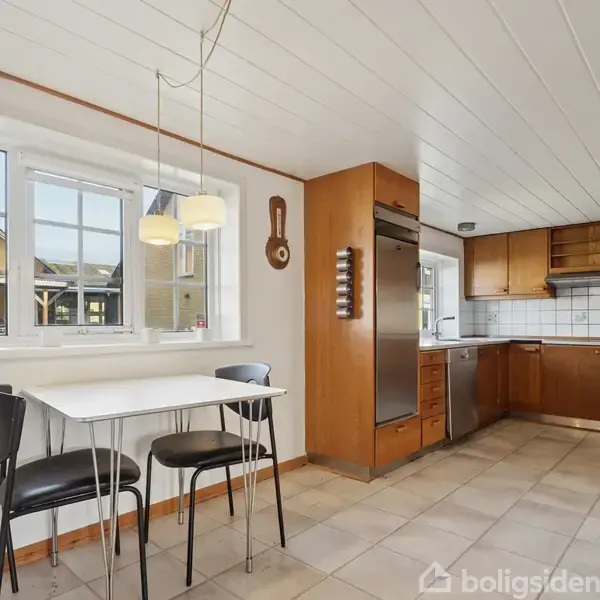 Et lyst køkken med hvide lofter og fliser. Et spisebord med to stole står foran vinduet. Brune skabe og hvide fliser pryder køkkenets bagvæg.