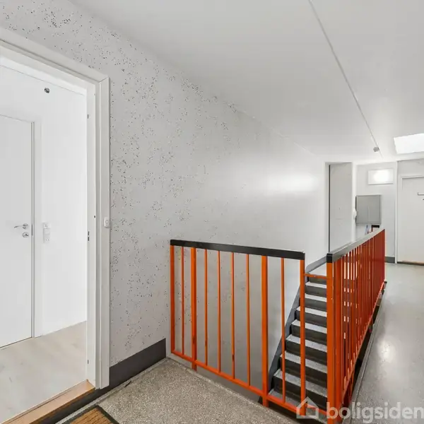 En lys trappeopgang med orange gelænder markerer en etageadskillelse. En åben hvid dør til venstre peger ind mod en lejlighed. Gulvene er grå og væggene hvide.