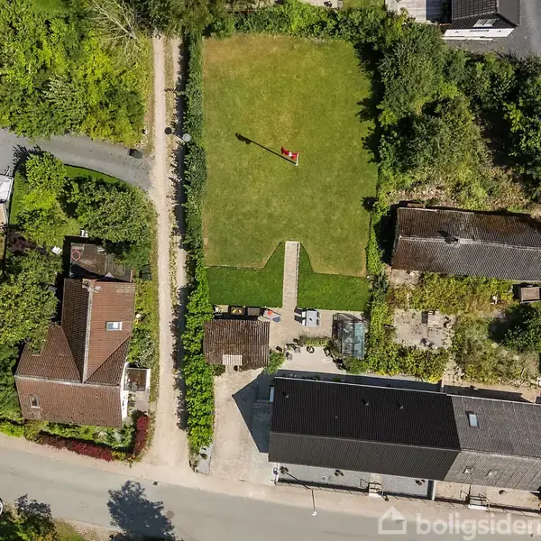 Grøn græsplæne med en rød flagstang i midten, omgivet af træer og buske. To huse med gårdspladser ligger på hver side af en vej i et boligområde.