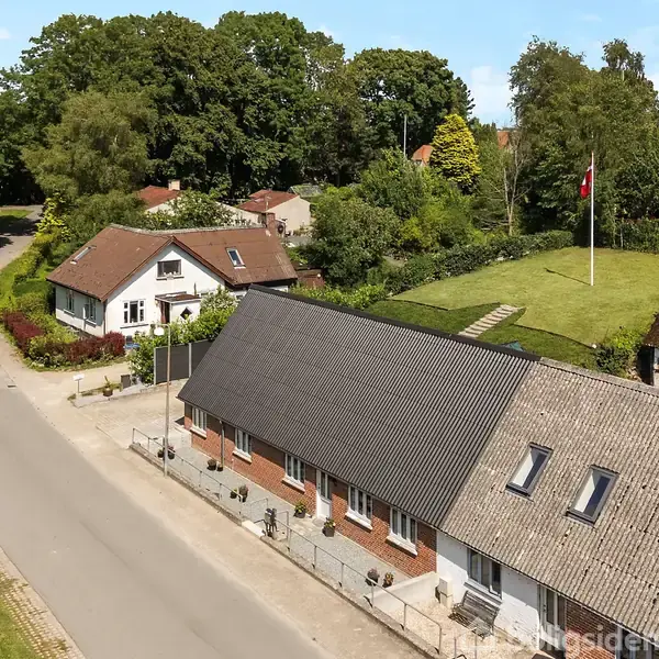En række huse med gavltage står langs en stille vej. En grøn græsplæne med flagstang ses i baggrunden. Omkringliggende træer skaber en rolig, landlig atmosfære.