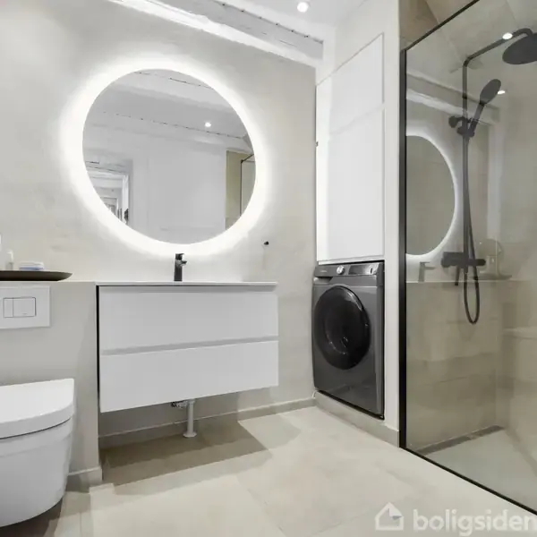 Et moderne badeværelse med væghængt toilet, rund spejl med lys, og vaskemaskine ved siden af en håndvask. Stor bruseniche med glasvæg til højre i et lyst, minimalistisk rum.