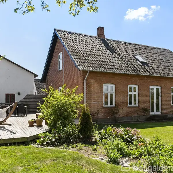 Rød murstenshus med mørkt tag står i en frodig have med blomsterbede. En træterrasse til venstre har en hængekøje og krukker, og en hvid bygning ses i baggrunden.