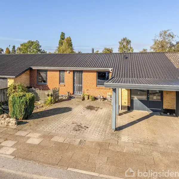 Et murstenshus med fladt tag, indkørsel og carport. Omgivet af en flisebelagt gårdsplads og små buske. Beliggende i et boligområde med flere liggende huse og træer i baggrunden.