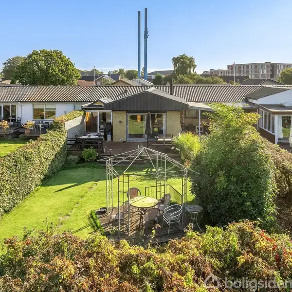 Et rækkehus med terrasseret have, hvoraf en del er indrettet som siddeområde med bord og stole. Hækken omkranser haven i et tæt beboelseskvarter med flere lignende huse.