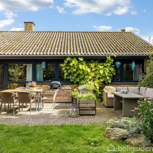 Huset har en terrasse med havemøbler og grill. En plante klatrer op ad facaden. Græsplæne omkranser terrassen, mens en blå himmel er synlig i baggrunden.