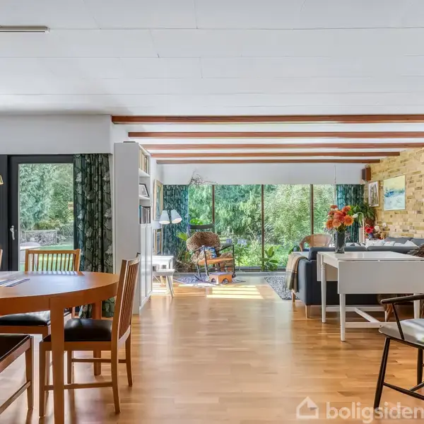 Spisestue med rundt træbord og stole, åben mod stueområde med sofaer. Store vinduer med udsigt til grøn have, lyserummet med naturligt lys. Trægulve og synlige loftsbjælker.