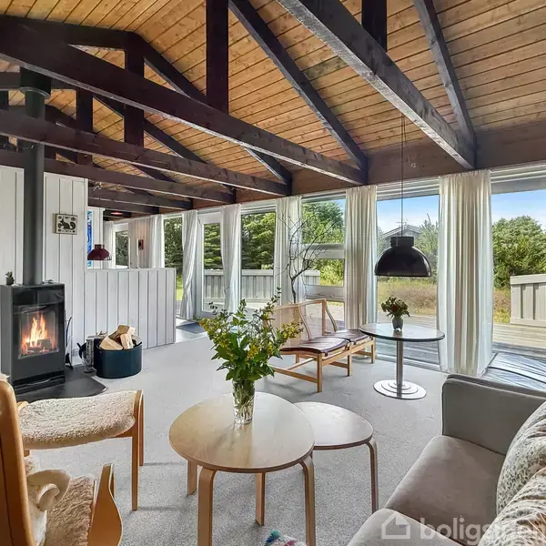 Stue med moderne møbler. Pejsen er tændt og blomster står på runde borde. Store vinduer giver udsigt til terrasse og grønne omgivelser. Træloft med synlige bjælker.