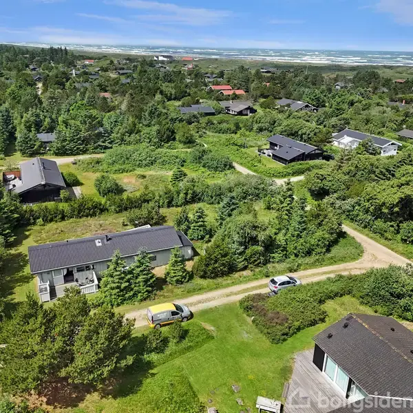 Et boligområde med spredte huse omgivet af frodig vegetation. To biler er parkeret på en grusvej mellem husene. Havet ses i baggrunden under en klar blå himmel.
