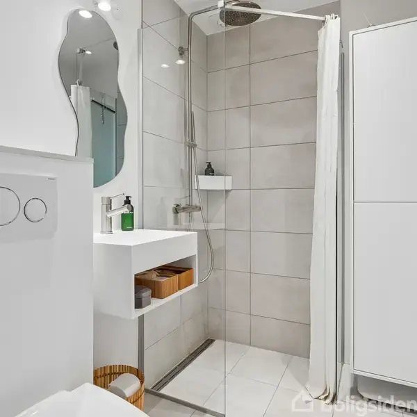 Et moderne badeværelse med hvidt sanitetsudstyr, hvor en bruser står i et hjørne med glasvæg og fliser. En vask står ved en væg, omgivet af opbevaring og spejl.