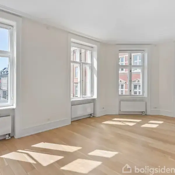 Bro strækker sig over en bykanal, omgivet af historiske bygninger og moderne højhuse. Grønne træer flankerer gaden, mens bådene flyder i vandet under en klar, blå himmel.