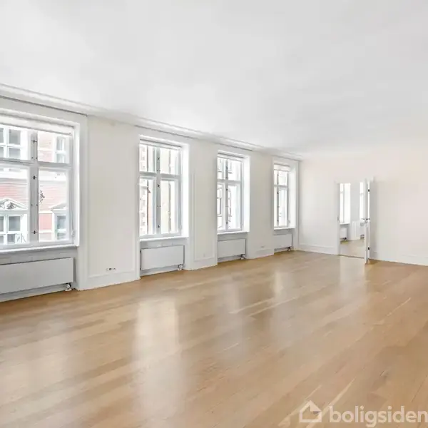 En rummelig, lys stue med poleret trægulv, fem store vinduer, og en moderne, hvid pejs i højre hjørne. Rummet er minimalistisk indrettet og har hvide vægge.