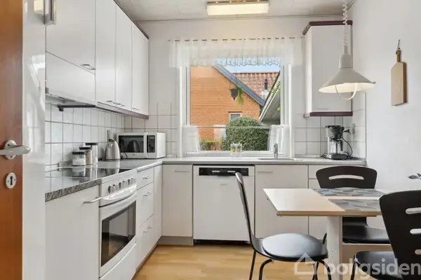 Et lyst køkken med hvide skabe og apparater, herunder en ovn, mikrobølgeovn og kaffemaskine. Et lille bord med to stole står ved vinduet med udsigt til en murstensbygning.