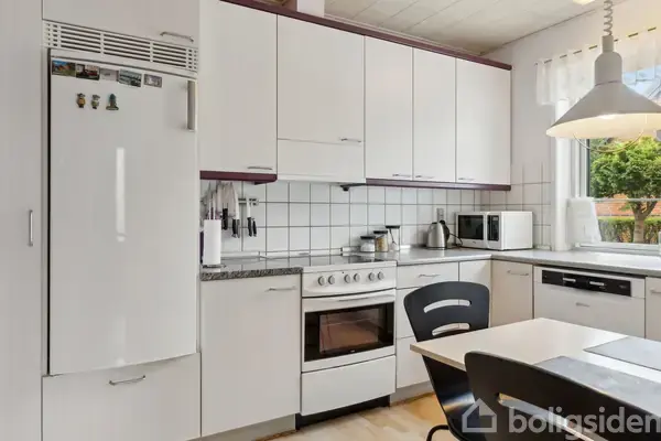 Et hvidt køkken med køleskab, komfur og ovn. Knive hænger på væggen. En mikrobølgeovn og kaffemaskine står nær et vindue med udsigt til træer.