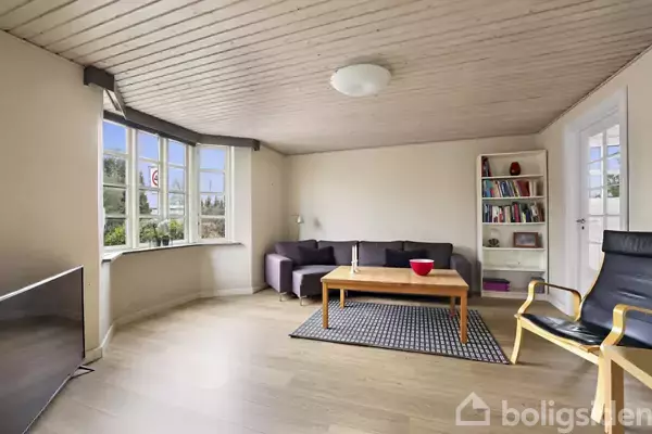En stue med en grå sofa står mod væggen. Et trækaffebord er foran sofaen, omgivet af en lænestol. Bøger fylder en reol ved siden af en lukket dør. Store vinduer til venstre giver lys.