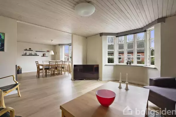 Stue med træbord, hvorpå en rød skål og to lysestager står. Fladskærms-tv ved siden af store vinduer. I baggrunden ses spisebord og stole under træloft.