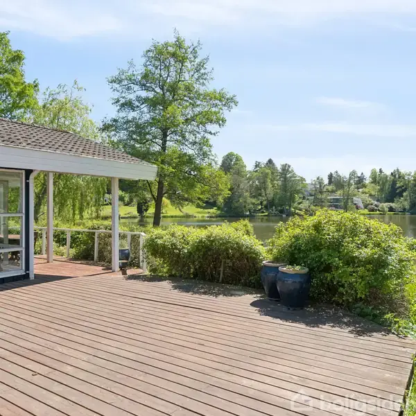 Terrasse med trædæk og et overdækket område ved siden af buske, der grænser op til en sø i en frodig, grøn have. Sollys skinner i en klar himmel.