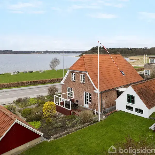 Et lyst spisestueområde med et rundt bord og seks stole i træ. Rummet har store vinduer med blomstrede gardiner, træloft og lyse trægulve, omkranset af skabe og planter.