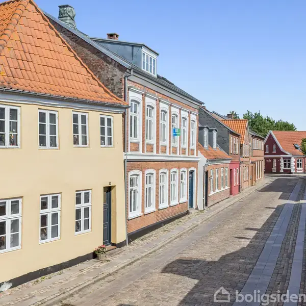 Et rækkehus med gule mursten og rødt tegltag står i en rolig, brolagt gade, flankeret af andre ældre huse. Solskin oplyser tækkeligt vedligeholdte facader.
