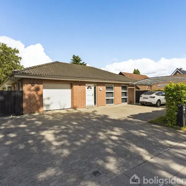 Rødt murstenshus med garageporte står stille på en flisebelagt indkørsel. En hvid bil er parkeret foran huset. Det er omgivet af grønne buske og områder med træer.