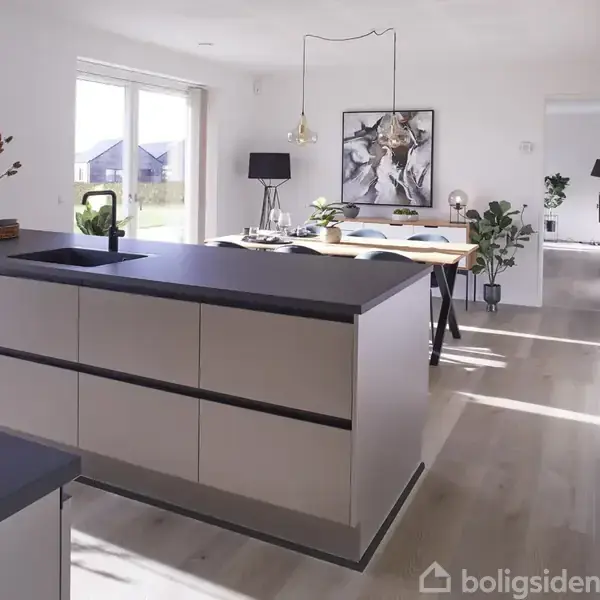 Et spisebord med blå stole står i et moderne køkken. Køkkenøen har planter og en ovn, mens store vinduer giver udsigt til grønne marker.